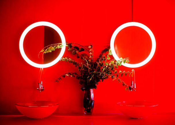 赤い色調のモダンなバスルームの写真、 2つの丸い形のスタイリッシュな洗面台、枝や葉を持つ透明な花瓶、コンクリート壁に小さなサイズの丸い照明ミラーのペア - 写真・画像