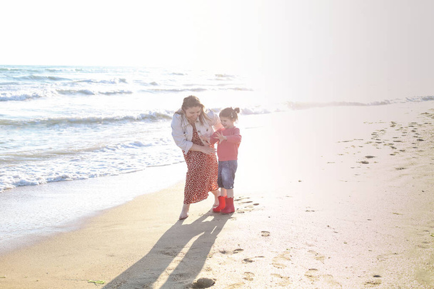 Glückliche zweiköpfige Familie, lächelnde Mutter und süße kleine Tochter, spazieren am sonnigen Sandstrand entlang, verbringen Zeit am Meer zusammen. Stimmungsvolles Lifestyle-Foto von aktiven Eltern, die mit Kindern unterwegs sind - Foto, Bild