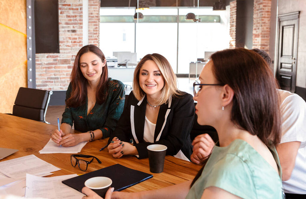 Groupe de jeunes professionnels du monde des affaires qui se réunissent dans une salle de réunion, quatre employés de bureau discutent ensemble d'un nouveau projet tout en étant assis sur un bureau dans un espace de coworking. Concept de travail d'équipe - Photo, image