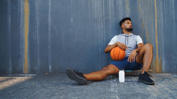 Young man with basketball doing exercise outdoors in city, sitting and resting. - Photo, Image