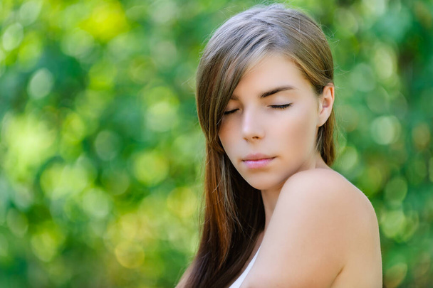 Μια νεαρή όμορφη ήσυχη γυναίκα με γυμνούς ώμους και κλειστά μάτια σε φόντο καλοκαιρινής πράσινης φύσης. - Φωτογραφία, εικόνα