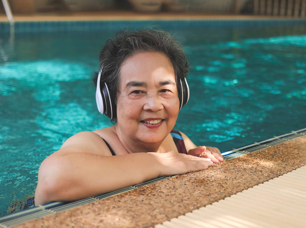 Muotokuva onnellinen ja terve aasialainen vanhempi nainen kuuntelee musiikkia kuulokkeet uima-altaassa, hymyillen ja katsoen kameraa. Iäkkäät aktiiviset elämäntavat. - Valokuva, kuva