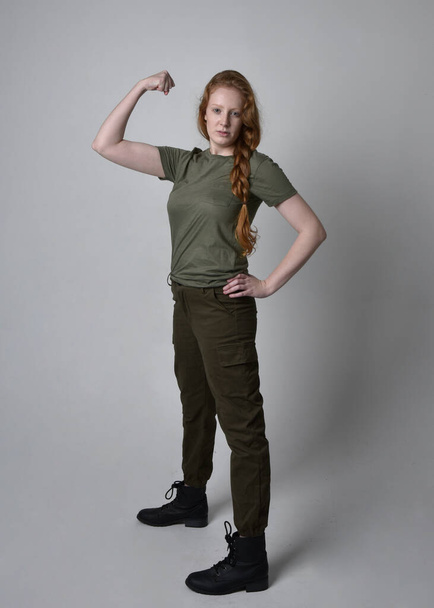 Portret pięknej rudej kobiety w wojskowej zielonej koszuli khaki, użytkowych spodniach i butach. Pozycja stojąca odizolowana na tle studia. - Zdjęcie, obraz