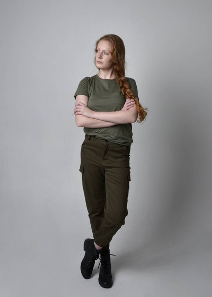 軍の緑のカーキシャツ、実用的なパンツやブーツを身に着けているかなり赤い髪の女性の完全な長さの肖像画。スタジオ背景に孤立した立ちポーズ. - 写真・画像