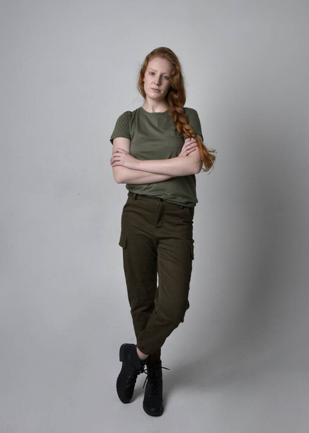 Full length portrait of pretty red haired woman wearing army green khaki shirt, utilitarian pants and boots. Standing pose isolated on studio background. - Photo, Image