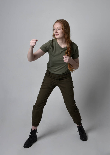 Uzun boylu, kızıl saçlı, ordu yeşili haki gömlekli, faydacı pantolon ve çizme giyen bir kadın portresi. Stüdyo arka planında izole edilmiş poz. - Fotoğraf, Görsel