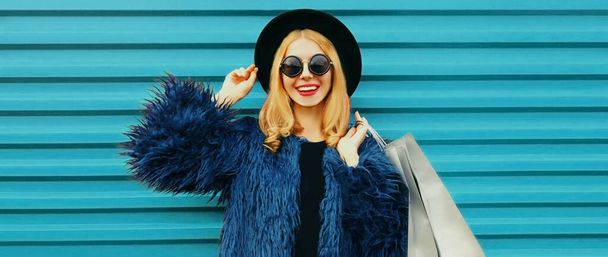Fashionable portrait of stylish blonde smiling woman with shopping bags wearing a blue faux fur coat, black round hat on colorful background - Photo, Image
