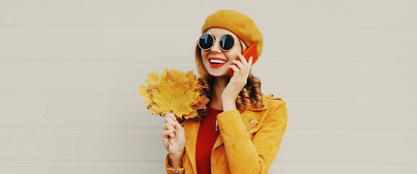 Autumn portrait of happy smiling young woman with yellow maple leaves calling on smartphone wearing a french beret over gray background - Fotó, kép