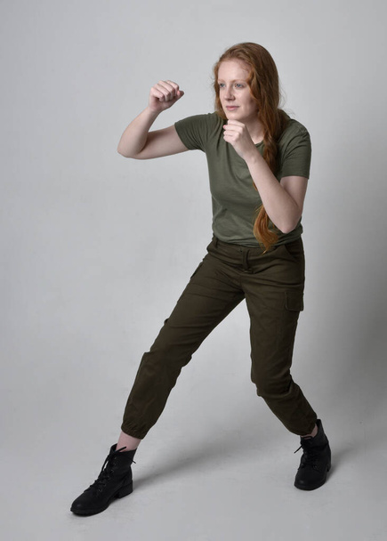Portrait complet d'une jolie femme aux cheveux rouges portant une chemise kaki vert armée, un pantalon utilitaire et des bottes. pose debout isolée sur fond de studio. - Photo, image