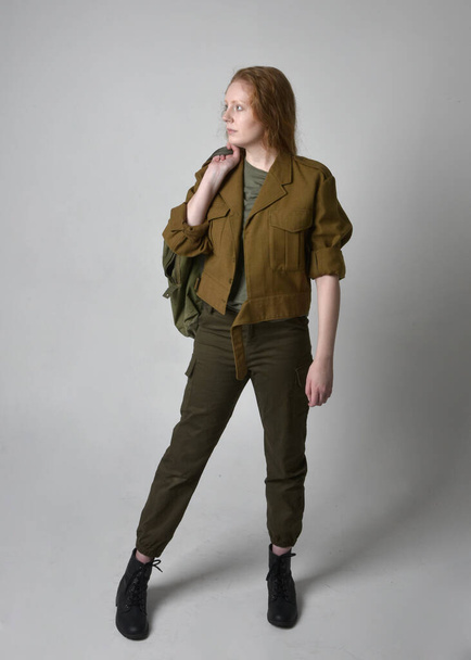 Full length portrait of pretty red haired woman wearing army green khaki shirt, utilitarian pants and boots. Standing pose isolated on studio background. - Photo, image
