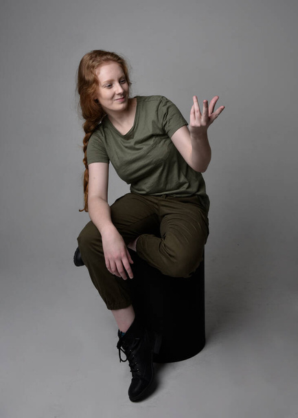 Retrato de cuerpo entero de una bonita mujer pelirroja vestida con una camisa verde caqui del ejército, pantalones utilitarios y botas. Posar sentado aislado en el fondo del estudio. - Foto, imagen