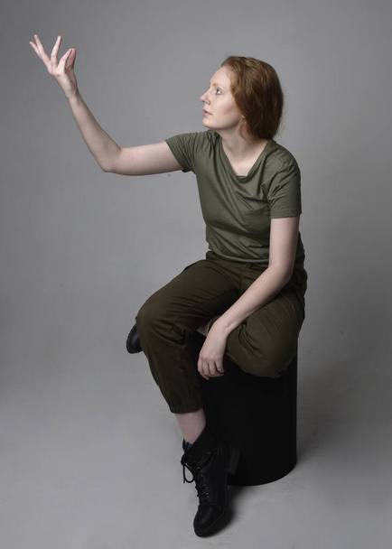 Portret pięknej rudej kobiety w wojskowej zielonej koszuli khaki, użytkowych spodniach i butach. Postawa siedząca odizolowana na tle studia. - Zdjęcie, obraz