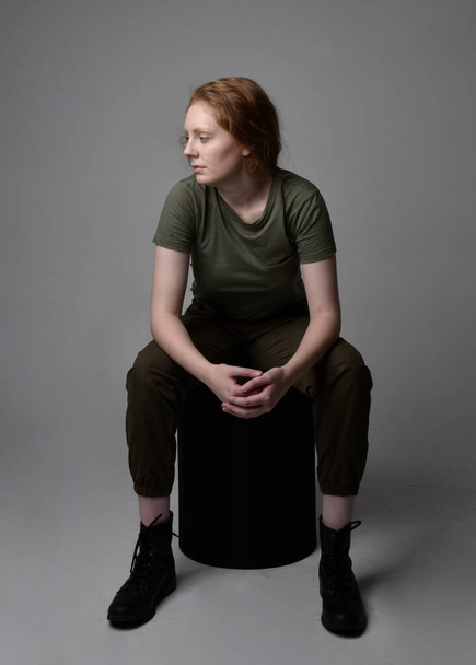 Retrato de cuerpo entero de una bonita mujer pelirroja vestida con una camisa verde caqui del ejército, pantalones utilitarios y botas. Posar sentado aislado en el fondo del estudio. - Foto, imagen