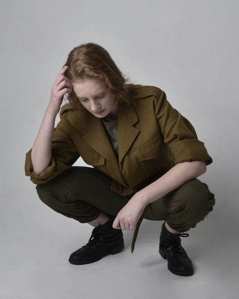Full length portrait of pretty red haired woman wearing army green khaki shirt, utilitarian pants and boots. Sitting pose isolated on studio background. - Fotoğraf, Görsel