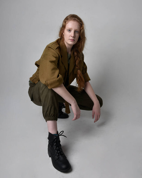 Full length portrait of pretty red haired woman wearing army green khaki shirt, utilitarian pants and boots. Sitting pose isolated on studio background. - Photo, Image