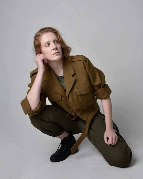 Full length portrait of pretty red haired woman wearing army green khaki shirt, utilitarian pants and boots. Sitting pose isolated on studio background. - Foto, Imagem