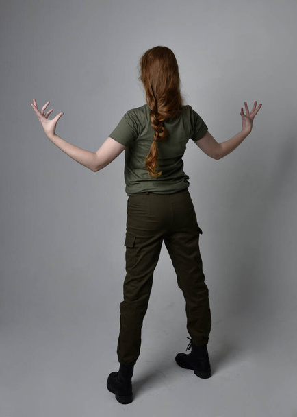 Full length portrait of pretty red haired woman wearing army green khaki shirt, utilitarian pants and boots. Standing pose with back to the camera, isolated on studio background. - Photo, Image