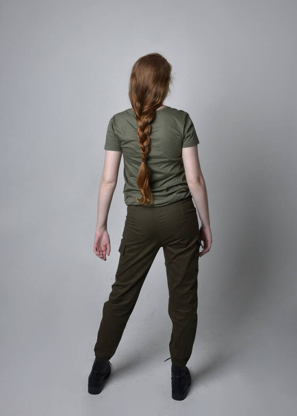 Retrato de cuerpo entero de una bonita mujer pelirroja vestida con una camisa verde caqui del ejército, pantalones utilitarios y botas. Posar de pie con la espalda a la cámara, aislado en el fondo del estudio. - Foto, imagen