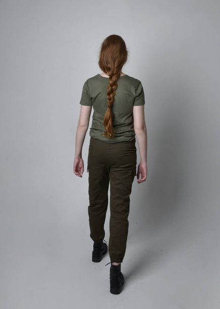 Portret pięknej rudej kobiety w wojskowej zielonej koszuli khaki, użytkowych spodniach i butach. Pozycja stojąca z tyłu do kamery, odizolowana na tle studia. - Zdjęcie, obraz