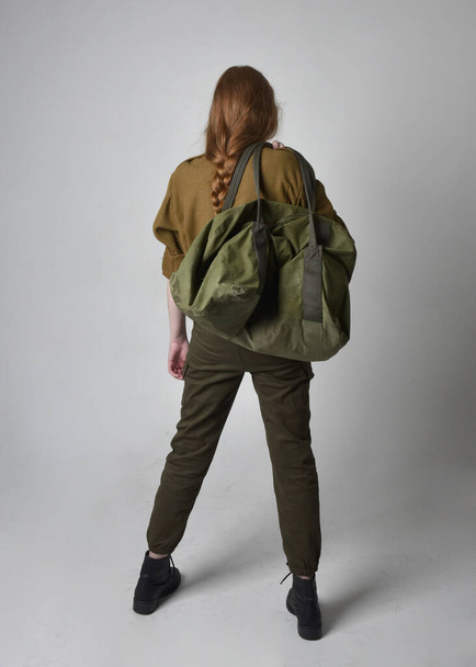 Plně dlouhý portrét krásné rudovlasé ženy v armádně zelené khaki košili, utilitárních kalhotách a botách. Stojící póza se zády ke kameře, izolované na pozadí studia. - Fotografie, Obrázek