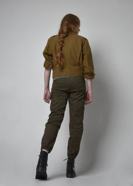 Полнометражный портрет рыжеволосой женщины в армейской зеленой рубашке хаки, утилитарных брюках и сапогах. Стоящая поза спиной к камере, изолированная на студийном фоне. - Фото, изображение