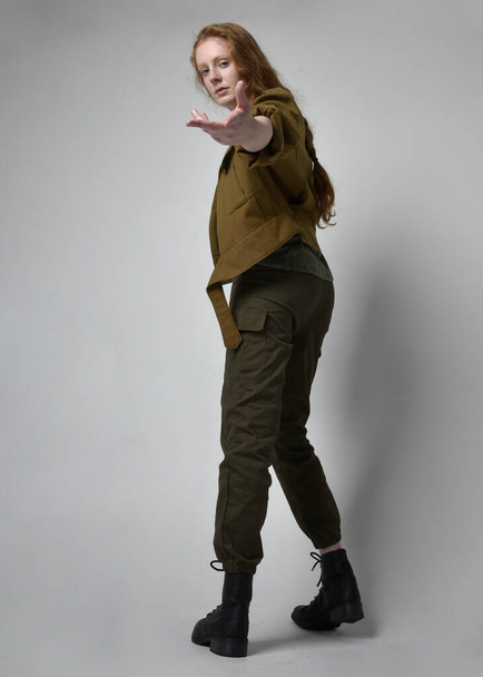 Full length portrait of pretty red haired woman wearing army green khaki shirt, utilitarian pants and boots. Standing pose with back to the camera, isolated on studio background. - Foto, imagen