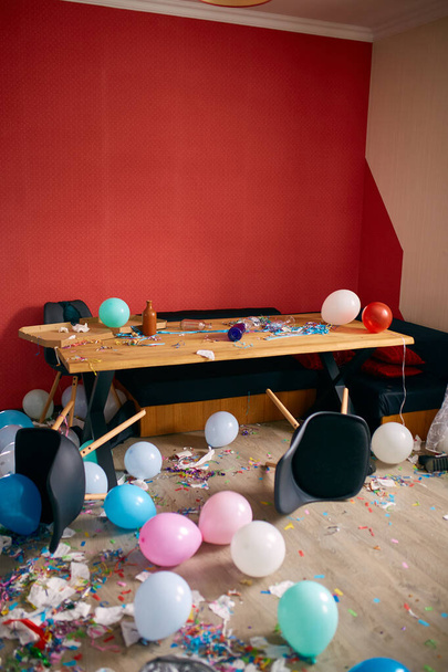 Nach dem Party-Chaos, chaotisch im heimischen Wohnzimmer, Tisch mit Pizza- und Sektgläsern, bedeckt mit Konfetti und Luftballons, Stuhl auf dem Boden am Morgen nach der Party-Feier. - Foto, Bild