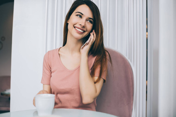 Молода приваблива жінка з чарівною посмішкою в повсякденній рожевій футболці, яка розмовляє по телефону, п'ючи чашку кави, сидячи в кафе, дивлячись далеко
 - Фото, зображення