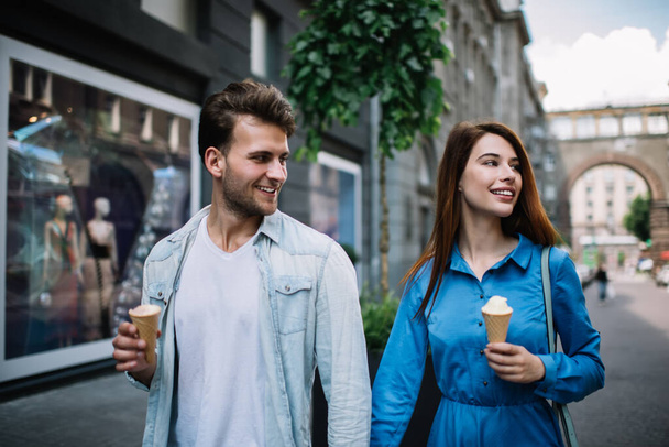 Ευτυχισμένο ρομαντικό ζευγάρι των εραστών σε casual ρούχα κοιτάζοντας μακριά και κρατώντας τα χέρια, ενώ απολαμβάνοντας το περπάτημα και το φαγητό παγωτό στο δρόμο της πόλης - Φωτογραφία, εικόνα