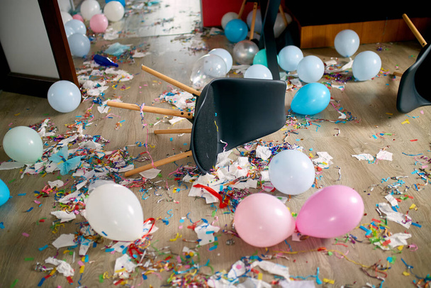 Po imprezie chaos, bałagan w salonie w domu, stół z pizzą i kieliszkami szampana pokrytymi konfetti i balonami, krzesło na podłodze rano po imprezie. - Zdjęcie, obraz