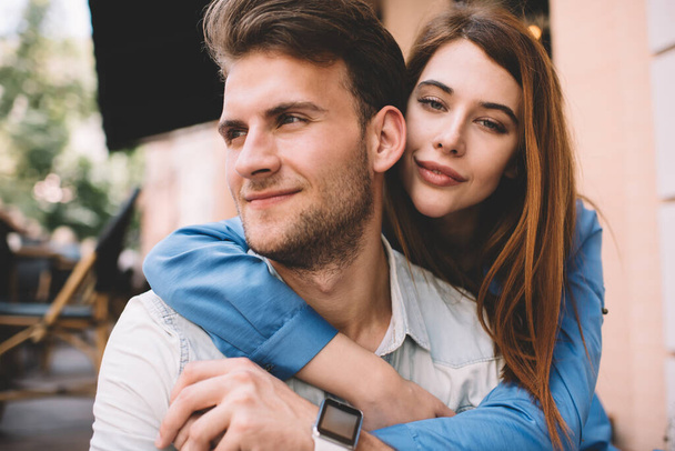 Beautiful woman looking at camera leaning on and hugging boyfriend form back while sitting together on terrace of cafe in summer time - Foto, Bild
