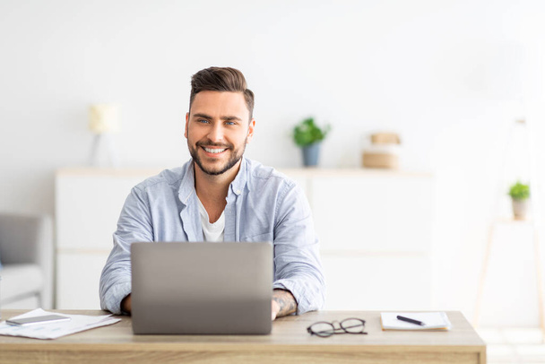 Úspěšná nezávislá osoba. Šťastný mladý muž sedí na pracovišti s notebookem a usmívá se do kamery, kopírovat prostor - Fotografie, Obrázek