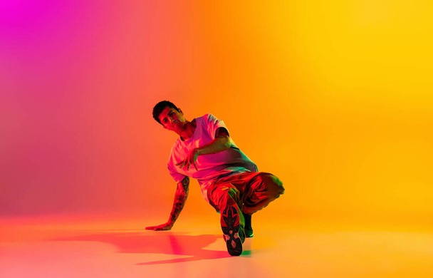 Full length portrait of young stylish man, break dancing dancer in casual clothes isolated over gradient pink yellow background at dance hall in neon light. - Fotografie, Obrázek