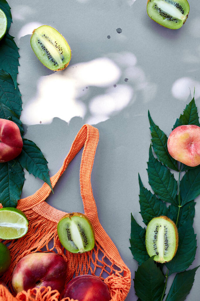 Плоский слой экологически чистой сумки для покупок с фруктами персиком, киви, лаймом на сером фоне при солнечном свете, летом. Продуктовая концепция, пространство для копирования, вид сверху. - Фото, изображение