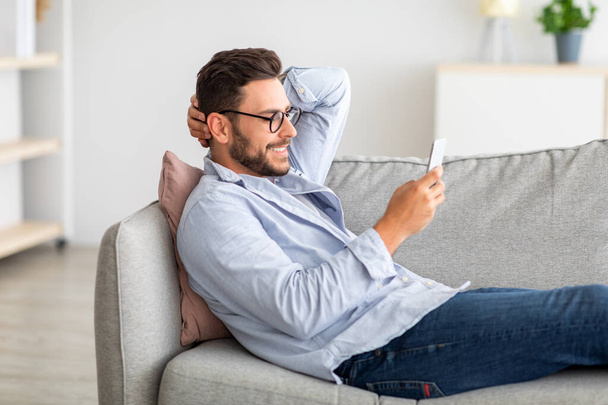 Cool gadget and app. Young smiling guy holding mobile phone, typing sms message, resting on sofa, free space - Photo, Image