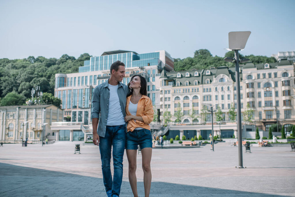 Content young traveling couple in casual wear walking in embrace and looking at each other against resort hotels in sunlight - Fotografie, Obrázek