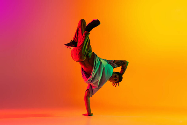 Portrait of young stylish man, break dancing dancer training in casual clothes isolated over gradient pink yellow background at dance hall in neon light. - Foto, afbeelding