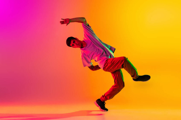 Portrait of young stylish man, break dancing dancer training in casual clothes isolated over gradient pink yellow background at dance hall in neon light. - Foto, Imagem