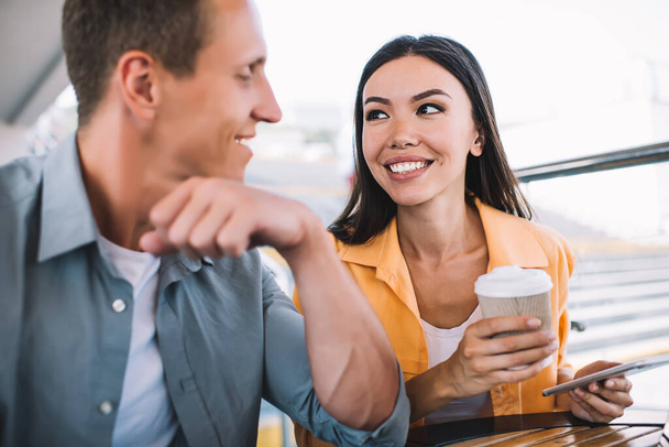 Relaxed smiling couple sitting at table together talking man leaning on hand and woman drinking coffee and using smartphone looking at each other - Foto, imagen