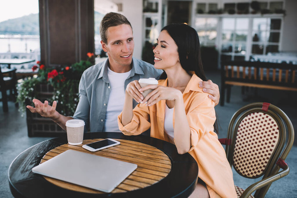 Улыбающаяся пара, сидящая за столом возле кафе с бумажными чашками кофе смартфона и ноутбука мужчина говорит и обнимает подругу слушать парня - Фото, изображение