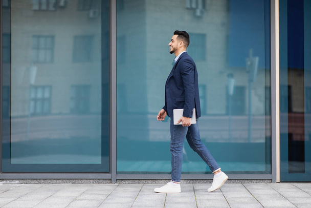 Cheerful arab businessman with pad walking by street, full length - Foto, Bild