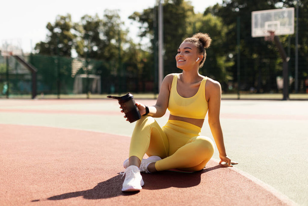 Porträt einer sportlichen schwarzen Frau in gelber Sportbekleidung, die Wasser trinkt - Foto, Bild