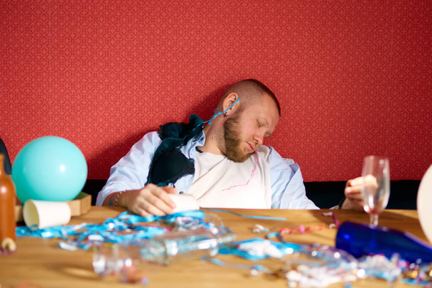 Homme barbu dormant à table dans une pièce désordonnée avec des sous-vêtements de filles, soutien-gorge après la fête de célibataire, homme fatigué après la fête à la maison - Photo, image