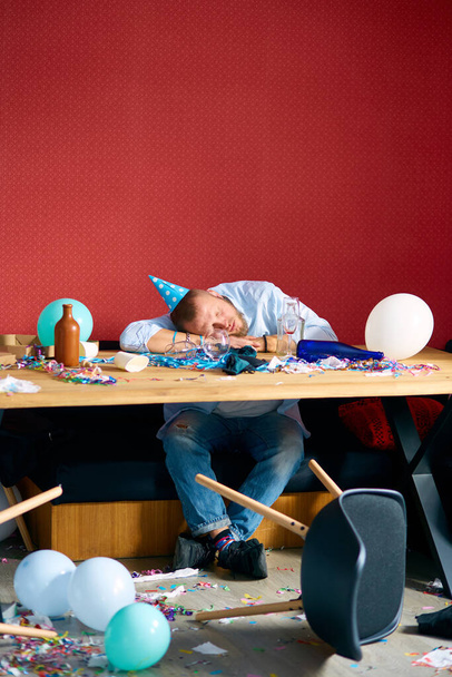 Man sleeping at table with blue cap in messy room after birthday party, tired man after party at home - Foto, afbeelding