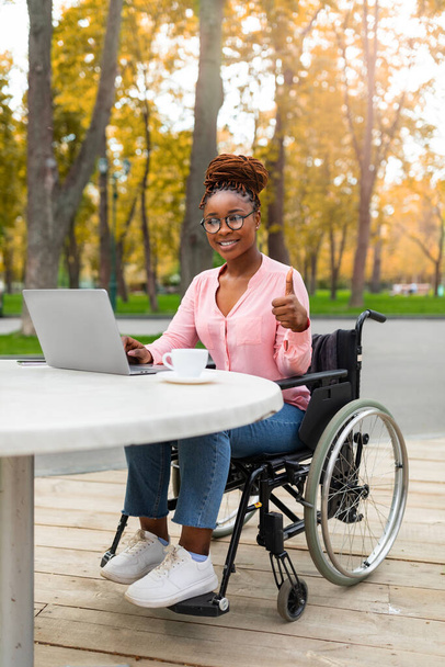 Dama negra discapacitada usando portátil, trabajando en línea, mostrando el pulgar hacia arriba en el parque de otoño. Trabajo remoto para minusválidos - Foto, imagen