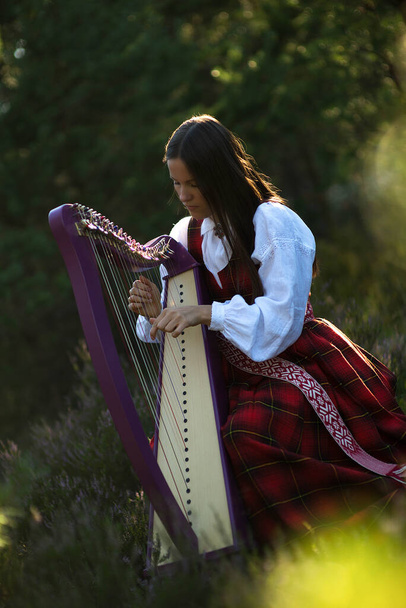 Portret kobiety z harfą.Piękna brązowłosa kobieta z wieńcem kwiatowym na głowie, ubrana w narodowy kostium grająca na harfie w lesie - Zdjęcie, obraz