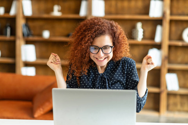 Overjoyed young curly woman surprised victory looks at the laptop - Photo, Image