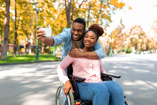 Cheerful black guy hugging his disabled girlfriend in wheelchair, making selfie on smartphone at autumn park - Photo, Image