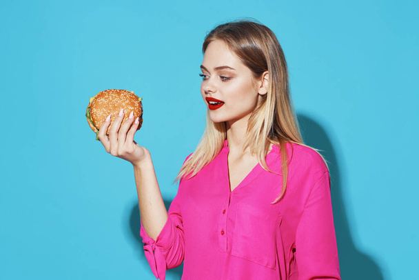 wesoła blondynka w różowej koszuli hamburger fast food snack niebieskie tło - Zdjęcie, obraz