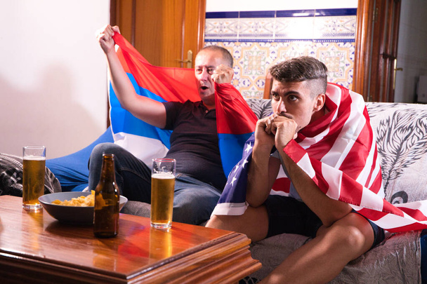 Dos amigos y fanáticos de los deportes están viendo un evento deportivo en la televisión mientras beben una cerveza. Cada uno de los hombres lleva una bandera sobre sus hombros. Concepto deportivo - Foto, imagen